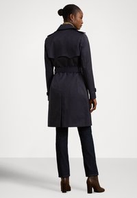 Σκούρο μπλε παλτό τύπου trench με ζώνη στη μέση, εμπρόσθιες πτυχώσεις στους ώμους και άνοιγμα στο πίσω μέρος, συνδυασμένο με σκούρο τζιν και καφέ μποτάκια με τακούνι.