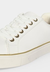 Witte lage sneaker met witte veters, gouden veteroogjes en een gouden streep langs de dikke rubberen zool.