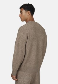 Młody mężczyzna stojący bokiem, ubrany w dopasowany jasnobrązowy sweter i spodnie na tle jasnej, jednolitej powierzchni.