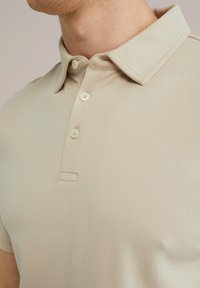 Camisa polo beige hecha de una tela suave, con cuello, tapeta de tres botones y detalles de costura sutiles.