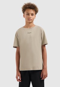 Tiener met krullend haar, gekleed in een beige t-shirt met korte mouwen met de tekst "BALLIN", staand tegen een effen lichtgrijze achtergrond.