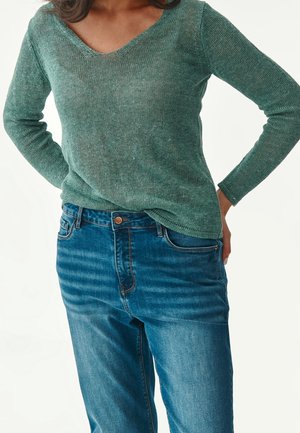Personne portant un pull en maille vert à manches longues et un jean bleu, les mains posées sur les hanches, sur un fond blanc uni.
