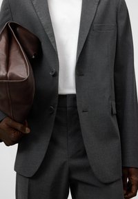 Completo grigio scuro con due bottoni, tasche laterali e rever a punta, tenuto con una mano. Nella altra mano è presente una borsa di pelle marrone scura.