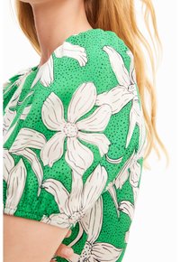 Zelená textilie s květinovým vzorem bílých květů s černými obrysy. Krátké, nariasené rukávy zvýrazňují texturu vzoru.