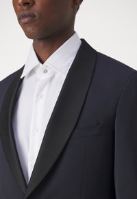 Schwarze Smokingjacke mit satinierter Revers, kombiniert mit einem weißen Hemd mit spitzem Kragen und Knopfleiste. Glatte Stofftexturen.