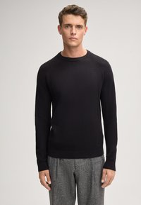 Zwarte gebreide trui met een textuurpatroon, ribbelkraag en boorden, met een getailleerde pasvorm. Gepaard met grijs geruite broek.