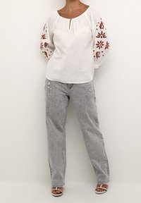 Personne portant une blouse blanche avec broderie florale rouge sur les manches, un jean clair déchiré et des sandales blanches à bout ouvert, se tenant contre un mur neutre.