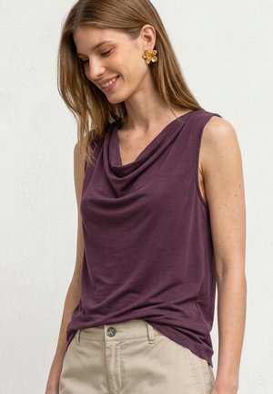 Femme aux cheveux bruns et lisses porte des boucles d'oreilles dorées en forme de fleur, un haut sans manches violet à col bénitier, et un pantalon beige, souriante et regardant vers le bas.