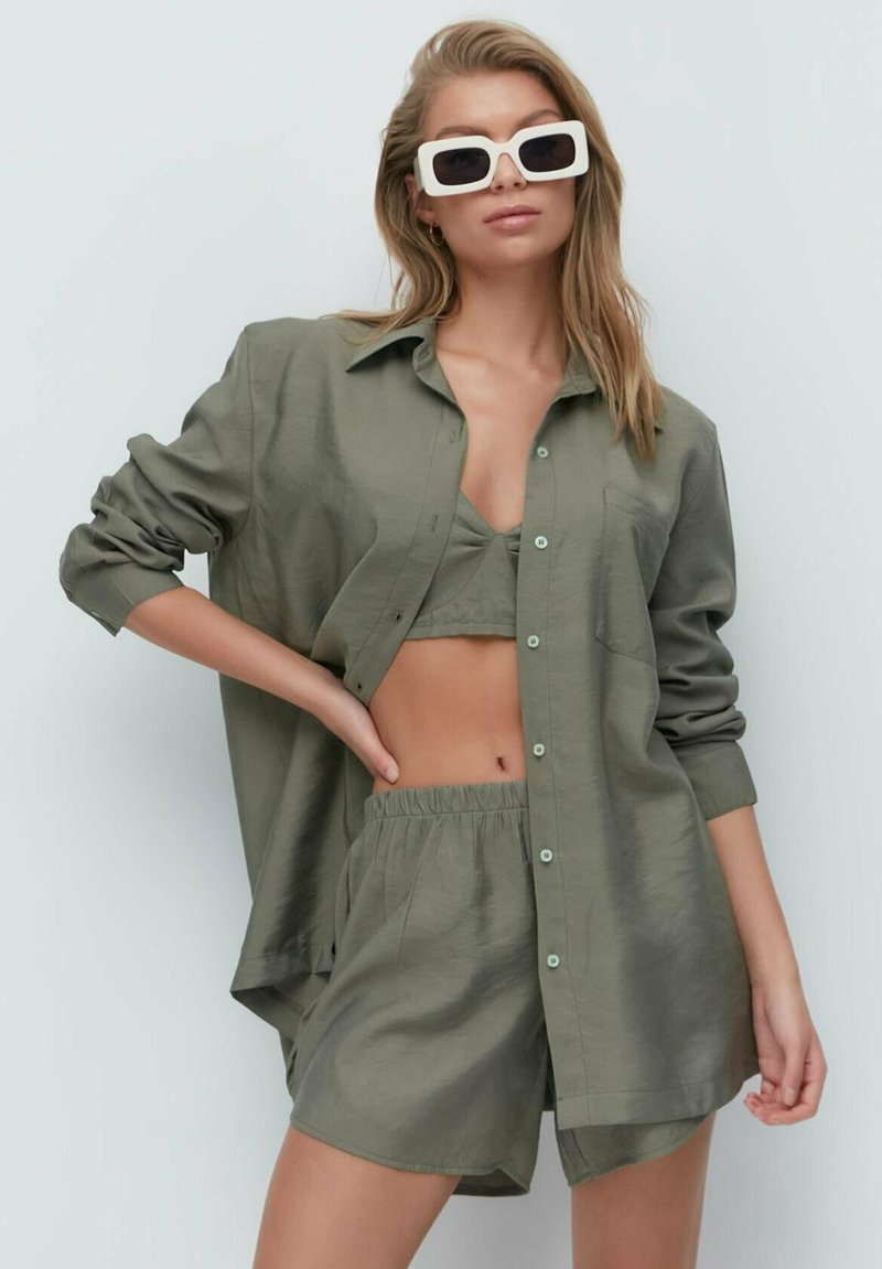 Olive green linen shirt with rolled sleeves, button closure, matching bikini top, and shorts; oversized fit; smooth texture. White rectangular sunglasses.