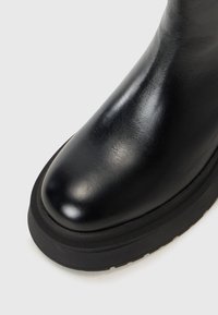 Gros plan sur une botte en cuir noir poli avec une épaisse semelle noire, mettant en valeur son design élégant et sa surface lisse.