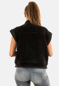 Svart ärmlös jacka med en strukturerad, mjuk krage och fåll. Buren över ljus denimjeans, vilket visar en avslappnad och ledig stil.