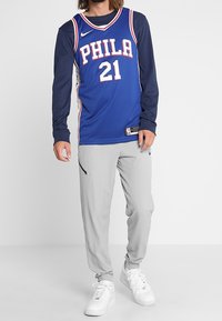 Uomo che indossa una maglietta da basket blu dei Philadelphia 76ers sopra una maglietta a maniche lunghe blu scuro, pantaloni grigi e sneakers bianche, in piedi contro uno sfondo neutro.