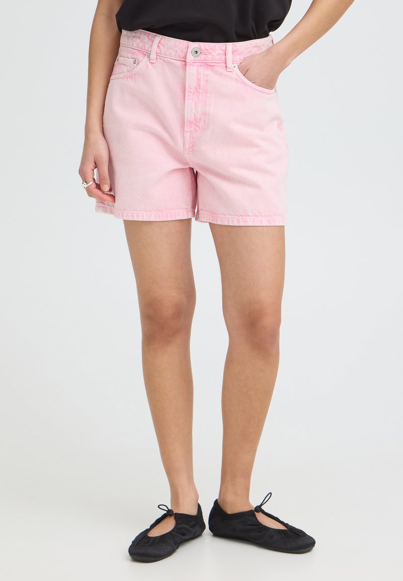 Personne portant un short en jean rose clair, un haut noir et des ballerines noires, debout avec une main dans la poche sur un fond uni.