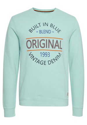 Gaiši zils džemperis, kas izgatavots no mīkstas auduma, ar apaļu kakla izgriezumu. Priekšpusē ir dizains ar tekstu "BUILT IN BLUE ORIGINAL VINTAGE DENIM 1993."