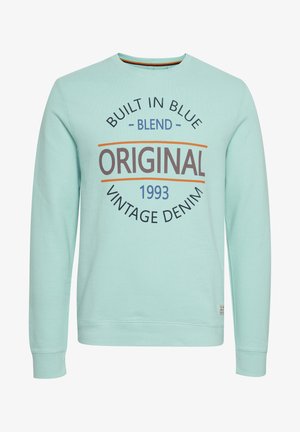 Gaiši zils džemperis, kas izgatavots no mīkstas auduma, ar apaļu kakla izgriezumu. Priekšpusē ir dizains ar tekstu "BUILT IN BLUE ORIGINAL VINTAGE DENIM 1993."