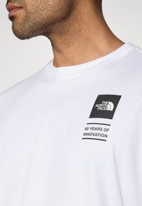 Λευκό βαμβακερό t-shirt με μαύρο τετράγωνο λογότυπο στο αριστερό στήθος, το κείμενο γράφει "60 ΧΡΟΝΙΑ ΚΑΙΝΟΤΟΜΙΑΣ" με μαύρα γράμματα.