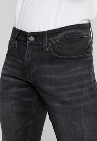 Mörkgrå denimjeans med en klassisk femfickdesign, subtila slitningar och en metallknappstängning vid midjan.