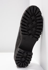 Semelle d’une botte en cuir noir avec un motif de bande de roulement profond et la taille 36 marquée, positionnée contre un mur blanc d’angle.