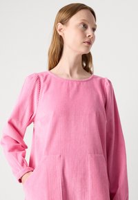 Ροζ βελουτέ τούνη με μακριά μανίκια, στρογγυλή λαιμόκοψη και δύο μπροστινές τσέπες. Το ύφασμα έχει κάθετη ριγωτή υφή.