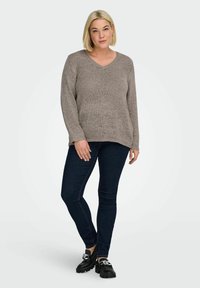 Blondýna stojící s rukou na stehně, oblečená v taupe úpletovém svetru, tmavých úzkých džínách a černých loaferech se stříbrnými řetízky.