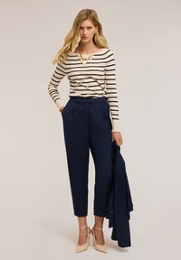 Una donna è in piedi indossando una maglia a righe crema e blu marino a maniche lunghe, pantaloni blu marino a vita alta, tacchi beige e tiene un blazer blu marino nella mano destra.
