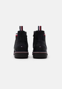 Mustat mokkanahkaiset nilkkurit, joissa on pieni korko, punavalkosinisävyinen vetotappi ja kontrastivalkoinen tikkaus pitkin pohjaa.