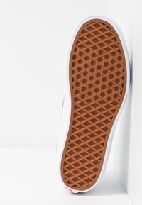 Gummisula på sneaker med ett brunt, strukturerat mönster med hexagonala och diamantformade figurer. Kanter i vitt med en lätt kurva och röd accent.