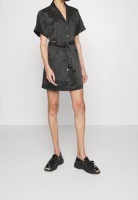 Robe noire à manches courtes avec boutons et détail de taille nouée, en tissu lisse ; portée avec des chaussures noires à lacets. Design minimaliste.