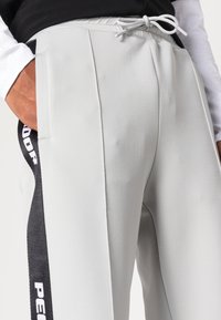 Ljust grå träningsbyxor med en slät yta, med sidofickor och svarta detaljer som visar vit text. Dragsko i midjan.