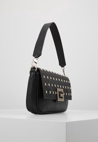 Sort lædertaske med en tekstureret overflade, nitter på klaffen, sølvhardware og en firkantet logoaccent. Kort, justerbar rem.