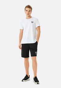 Hvid kortærmet t-shirt med lille sort tryk på brystet, parret med sorte shorts med hvid "TRAINING 98" tekst. Sorte sportssko.