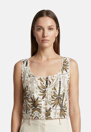 Woman with straight brown hair wears sleeveless white crop top with brown and green leaf patterns and scalloped edges, paired with beige pants.