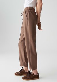 Personne portant un pantalon brun ajusté jusqu'aux chevilles avec taille élastique et des baskets brunes, se tenant avec les mains croisées derrière le dos.