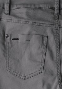 Pantaloni in tessuto grigio con una tasca posteriore caratterizzata da cuciture in contrasto e un bottone metallico. Include un piccolo etichetta del marchio vicino alla tasca.
