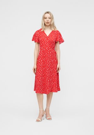 Lyshåret kvinde iført en rød kjole til knæet med hvide blomsterprint og flagrende ærmer, kombineret med hvide sandaler med høje hæle.