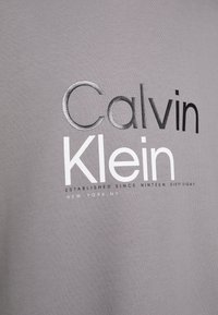 T-shirt in cotone grigio con il nome del marchio "Calvin Klein" in lettere in grassetto nero, grigio e bianco, con una finitura testurizzata.