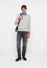 Grau gestrickter Pullover mit V-Ausschnitt, darunter ein Denim-Hemd, dunkelgraue Jeans und schwarze Slipper. Zubehör umfasst eine bordeauxfarbene Mütze und eine schwarze Tasche.
