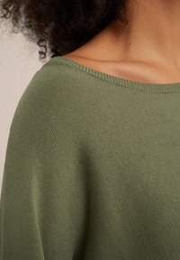 Pull en maille verte avec un large col et une texture côtelée, mettant en valeur un tissu doux et une coupe oversize.