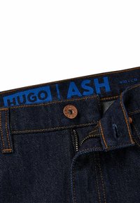 Jeans de mezclilla azul oscuro con costuras naranjas, que cuentan con un botón de cobre y un cierre de cremallera. La etiqueta dice "HUGO | ASH" en texto azul en negrita.