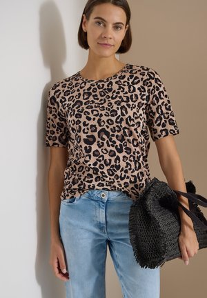 Femme portant un haut à manches courtes imprimé léopard et un jean bleu clair, tenant un sac noir tissé sur un fond neutre.