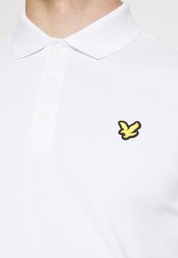 Valkoinen polo-paita, jossa on kolmipainikkeinen kaulus ja musta-keltainen lintulogo vasemmassa rinnassa. Sileä pinta, puuvillasekoitemateriaali.