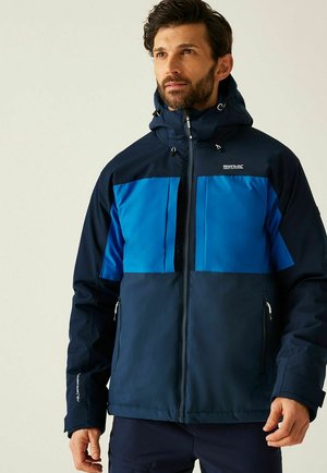 Hombre con barba que lleva una chaqueta de exterior con capucha en tonos azul marino y azul, con cremallera frontal y bolsillos con cremallera, mirando ligeramente hacia arriba y a la derecha.