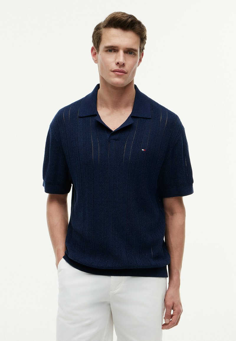 Young man wearing a navy blue textured short-sleeve polo shirt and white pants, standing against a plain white background.