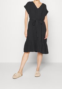 Vestido negro con manga corta, escote en V y una cintura ceñida con lazo, que presenta un dobladillo acampanado. Combinado con zapatos beige sin cordones.