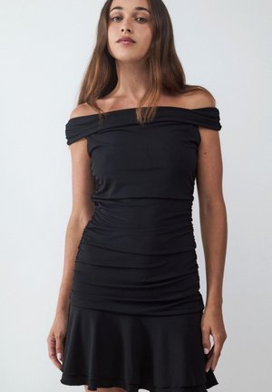 Jeune femme aux longs cheveux bruns portant une robe noire à épaules dénudées et froncée, debout devant un fond clair uni.