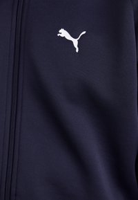 Mørkeblå teksturert tracksuit-jakke med full glidelås, med en hvit Puma-logo på øvre venstre brystområde.