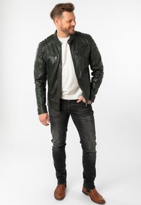 Dunkelgrüne Lederjacke mit gesteppten Schulterakzenten, zwei mit Reißverschluss versehenen Brusttaschen, getragen über einem weißen Hemd und kombiniert mit dunkelblauen Jeans und braunen Stiefeln.