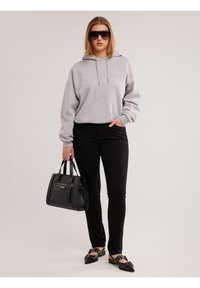 Mujer con sudadera gris con capucha, pantalones negros, zapatos negros con tachuelas, grandes gafas de sol y bolso negro, de pie contra un fondo liso.