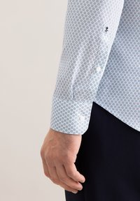 Hellblaues Hemd mit geometrischem Muster, weißen Knöpfen und Manschettendetail. Der Stoff wirkt glatt mit einer eleganten Oberfläche.