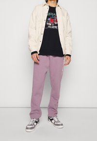 Chaqueta crema de botones sobre una camiseta gráfica negra, combinada con pantalones de chándal suaves y morados claros y zapatillas gris-negras.